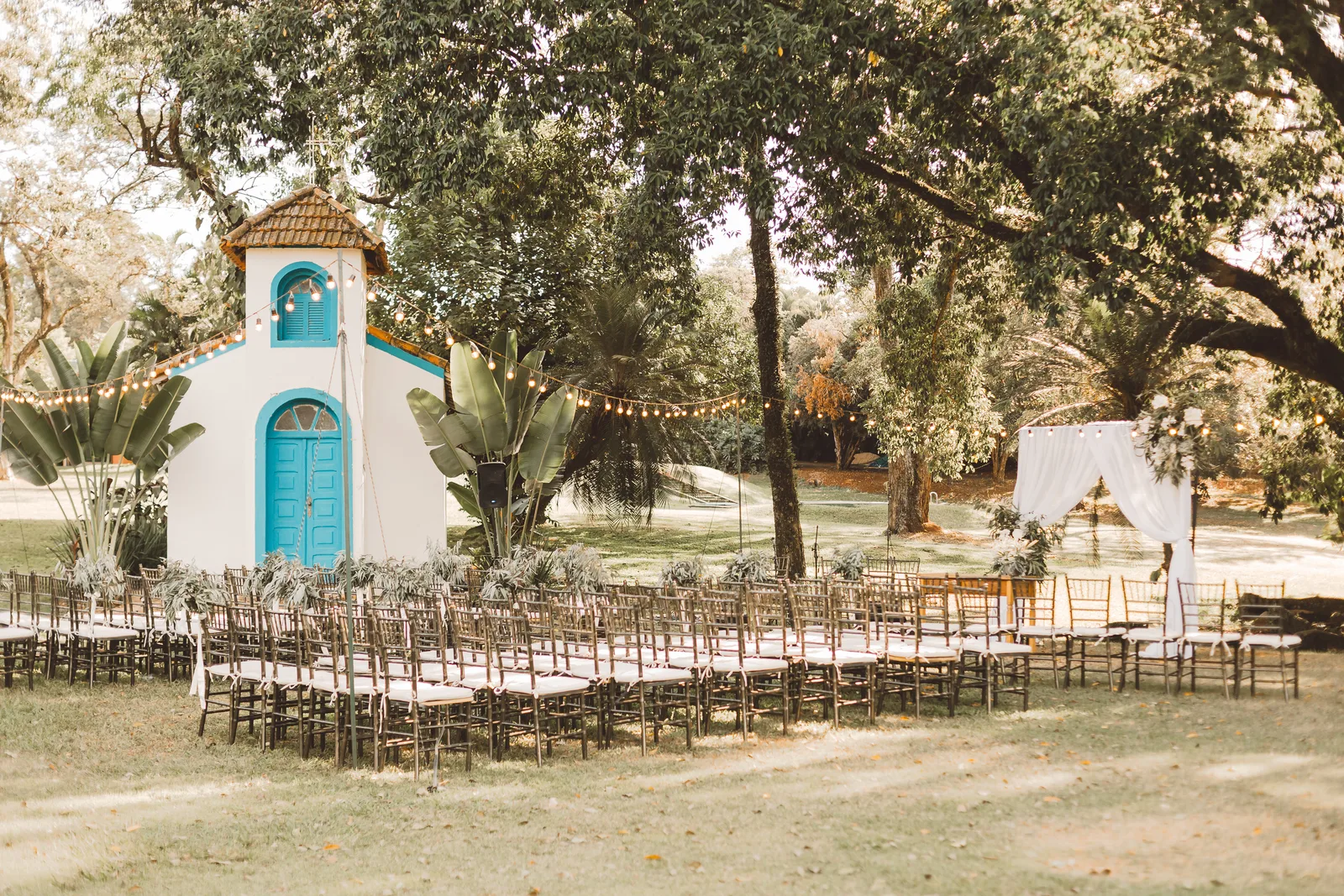 Cadeiras da cerimônia sob as árvores da Fazenda A Querência, ao lado de uma pequena capela branca de porta azul